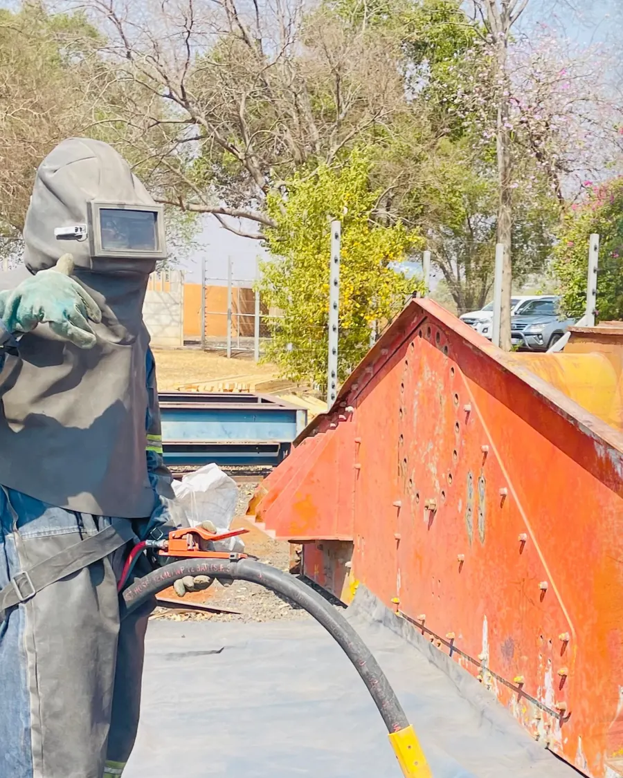 Rusted orange steel chute before sandblasting, operator holding blast hose
